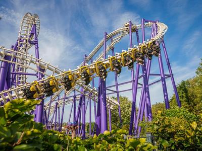 Roller coaster z fioletowymi podporami i żółtymi wagonikami jedzie przez zielone liście pod niebieskim niebem.