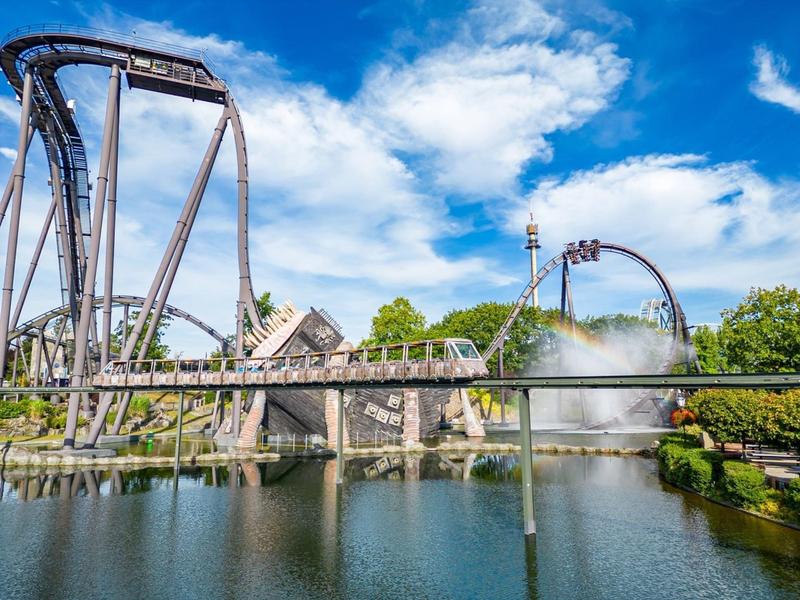 Rollercoaster przejeżdża przez most nad wodą pod niebieskim niebem w parku rozrywki.
