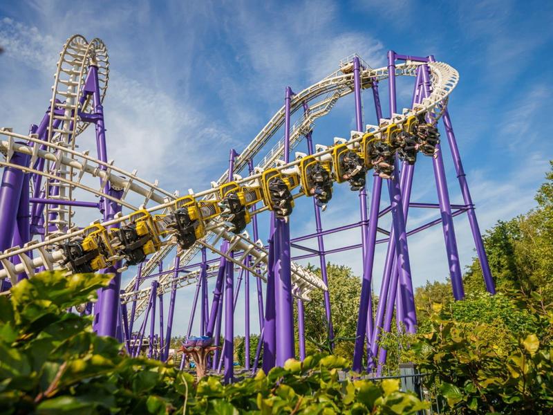 Roller coaster z fioletowymi podporami i żółtymi wagonikami jedzie przez zielone liście pod niebieskim niebem.
