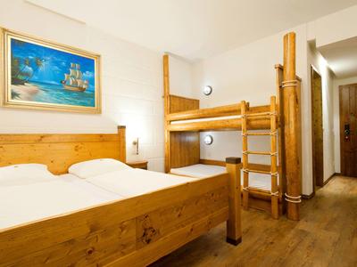 Hell eingerichtetes Hotelzimmer mit Doppelbett und Etagenbett aus Holz, maritimes Bild an der Wand.