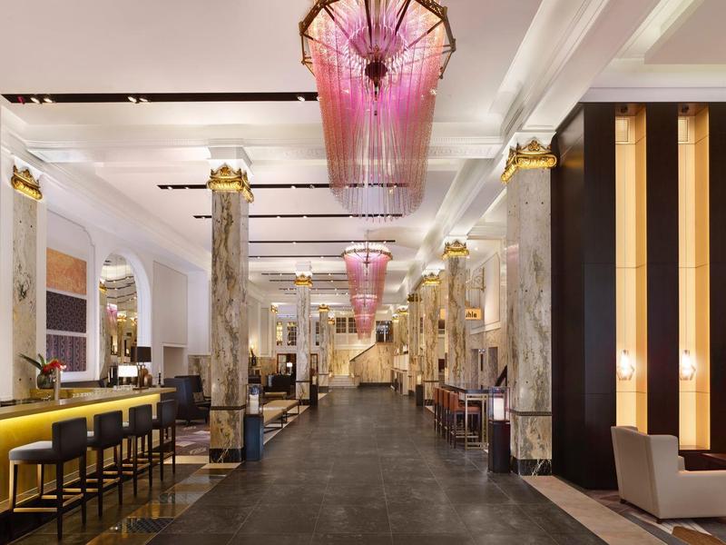 Moderne Hotel-Lobby mit langer Bar, hohen Decken, Säulen und pinken Lichtinstallationen.