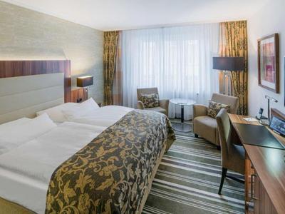 Helles Hotelzimmer mit großem Bett, zwei Sesseln, Schreibtisch und Fenster mit weißen Vorhängen.