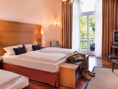 Helles Hotelzimmer mit Doppelbett, Stuhl, Fernseher und Balkon mit Blick ins Grüne.