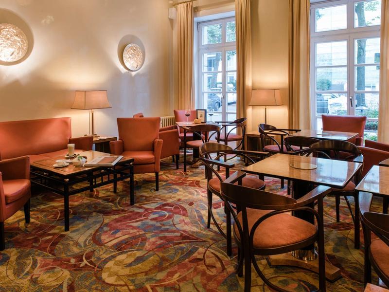 Gemütlicher Caféraum mit roten Sesseln, Holztischen, Teppichboden und großen Fenstern.