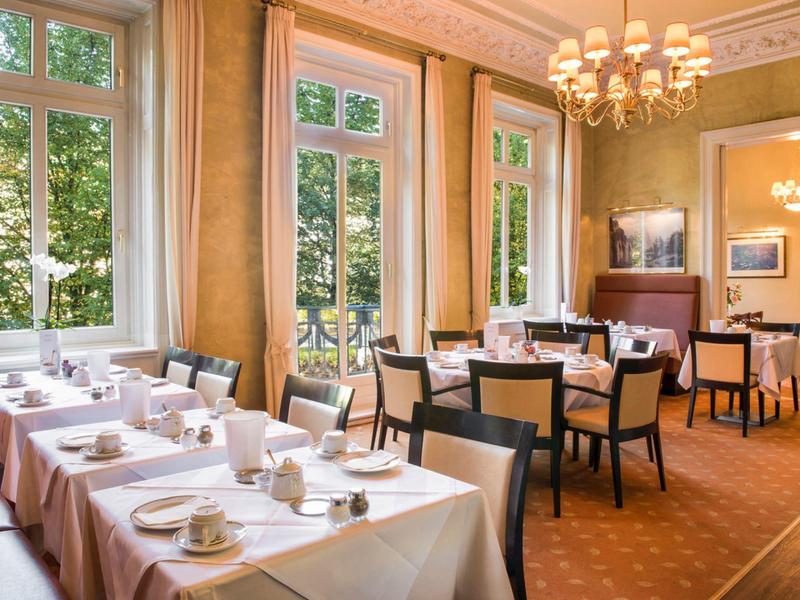 Helles Restaurant mit großen Fenstern, weißen Tischdecken, dunklen Stühlen und Kronleuchtern.