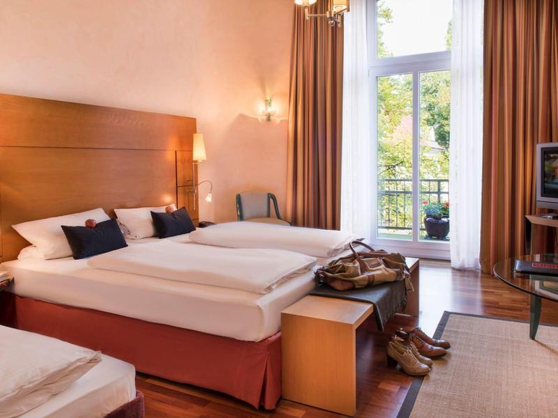 Modernes Hotelzimmer mit zwei Betten, Holzmöbeln, Balkon und Fernseher. Helle, warme Farben.