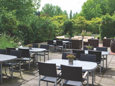 Leerer Außenbereich eines Cafés mit Tischen und Stühlen, umgeben von grünen Bäumen.