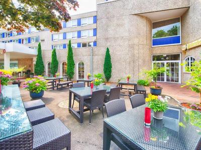 Moderner Außenbereich eines Hotels mit bepflanzten Tischen und Stühlen unter Bäumen.