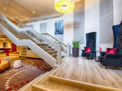 Moderner Hotel-Eingangsbereich mit Treppe, Sesseln und Teppichboden in warmen Farben.