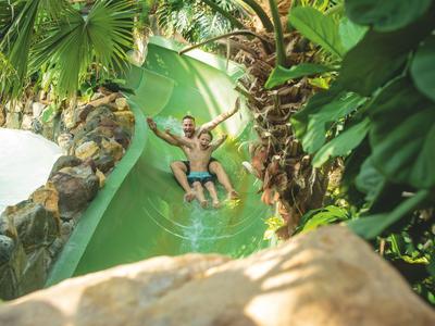 Kind rutscht fröhlich auf einer grünen Wasserrutsche in tropischer Umgebung