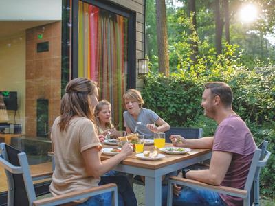 Familie sitzt draußen beim Frühstück in einem Garten mit modernem Haus im Hintergrund.