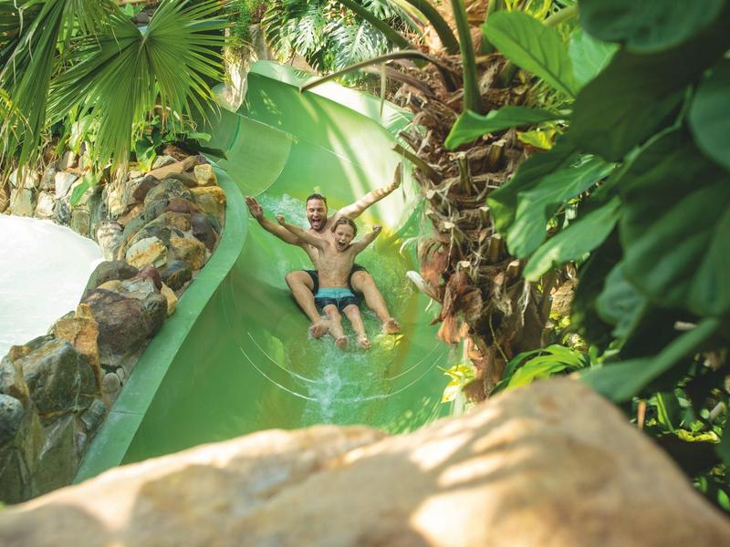 Muž sjíždí zelenou vodní skluzavku v tropickém vodním parku.