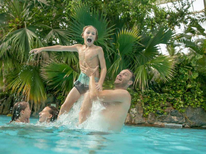 Otec zvedá dítě v bazénu obklopeném bujnými tropickými rostlinami.