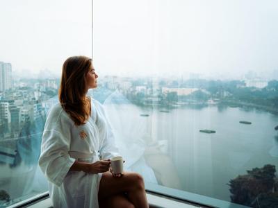Frau im weißen Bademantel sitzt am Fenster und blickt auf einen Fluss mit Booten in der Stadt.