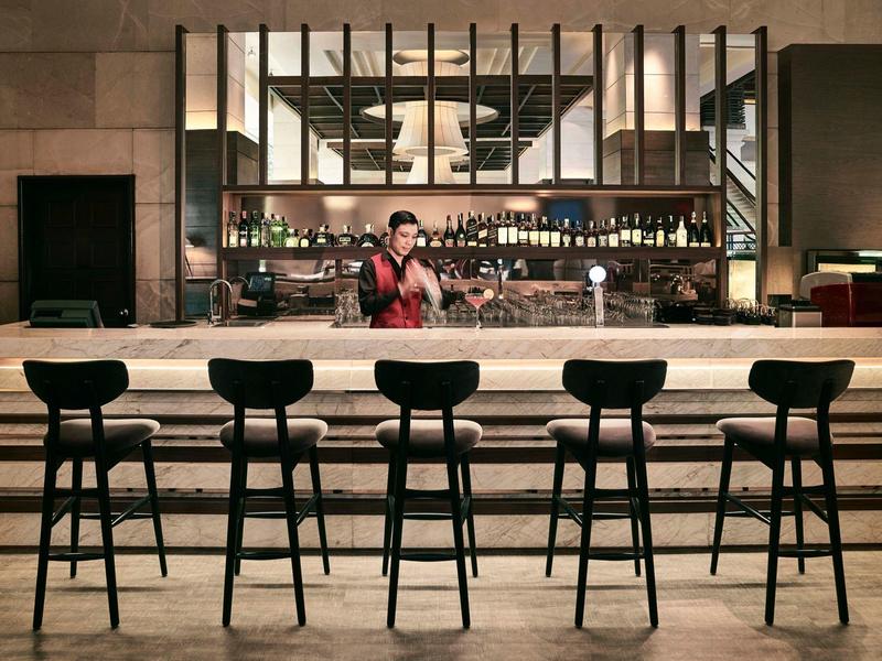 Moderne Bar mit fünf schwarzen Barhockern und Barkeeper hinter langer Theke aus Holz und Marmor.