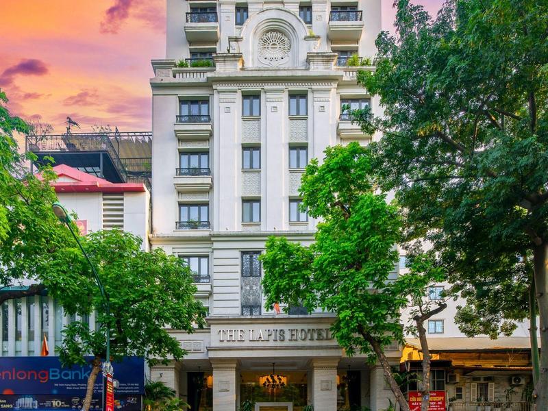 Elegant hotelgebouw tegen een kleurrijke zonsondergang met bomen en straat ervoor.