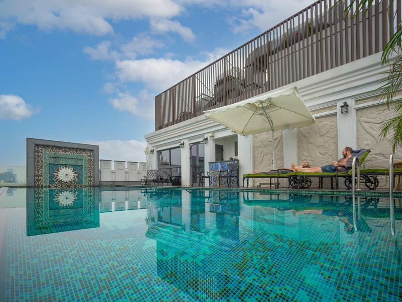 Dakterraszwembad met blauwe tegels, ligbedden en zonnescherm onder deels bewolkte blauwe lucht.