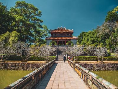 Ein Weg führt zu einem traditionellen asiatischen Gebäude, umgeben von Bäumen und klarem Himmel.