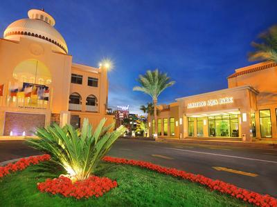 Bâtiment d'hôtel moderne avec façade éclairée, palmiers et jardin entretenu au crépuscule.