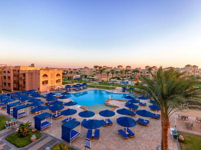 Grande zone de piscine avec parasols bleus entourée de bâtiments d'hôtel au lever du soleil.