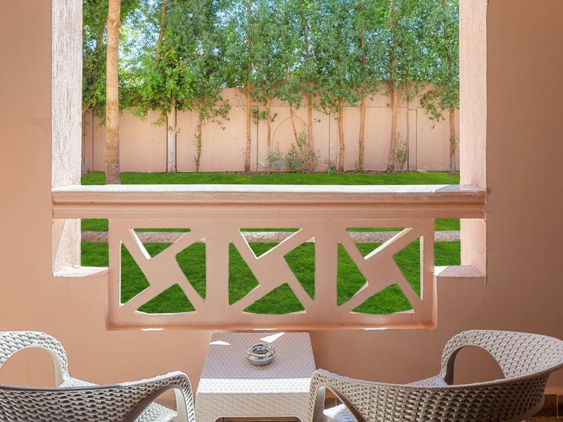 Zwei weiße Stühle und ein kleiner Tisch auf einer Terrasse mit Blick auf grüne Bäume hinter einer rosa Wand.