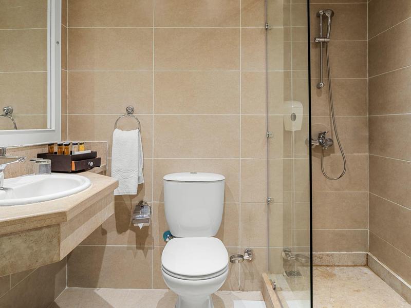 Kompaktes Badezimmer mit weißer Toilette, eckigem Waschbecken, Duschecke und hellbeigen Fliesen.