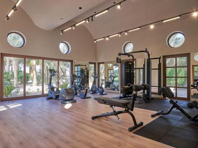 Bright gym with wooden floor, various equipment, and large windows overlooking palm trees.