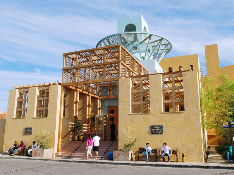 Nowoczesny budynek z drewnianym tarasem i fontanną, jasno niebieskie niebo, ludzie siedzą i stoją.