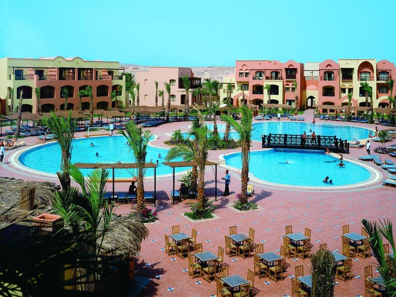 Duży kompleks hotelowy z okrągłym basenem, palmami i leżakami.