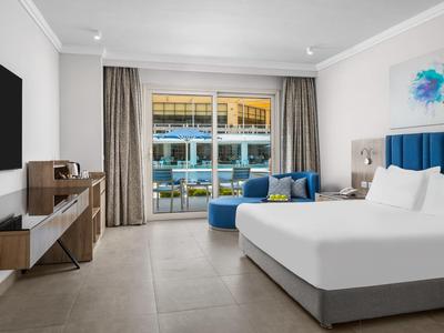 Camera d'hotel moderna con letto matrimoniale, poltrona blu e balcone con vista piscina.