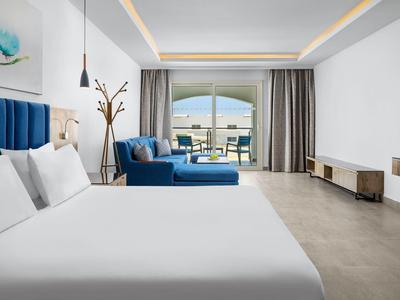 Camera d'hotel moderna con letto grande, poltrona blu e vista sul balcone soleggiato.