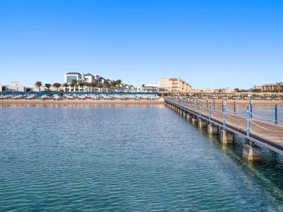 Lange pier over blauwe zee met hotels en palmbomen op de achtergrond onder een heldere lucht.