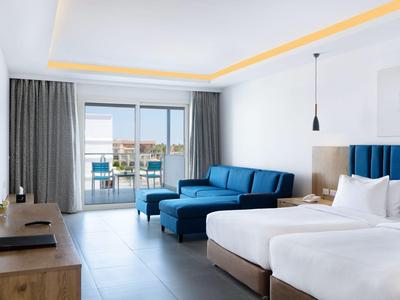 Modern hotelkamer met kingsize bed, blauwe bank, balkon en zeezicht.