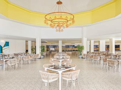 Ampia sala da pranzo luminosa con tavoli rotondi e un lampadario sotto una cupola gialla.