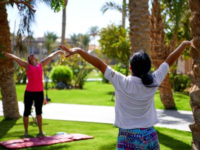 Two women doing yoga outdoors on green grass with palm trees in the background.