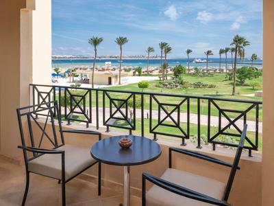 Balcony with table and chairs overlooking palm trees, beach, and sea under a blue sky.