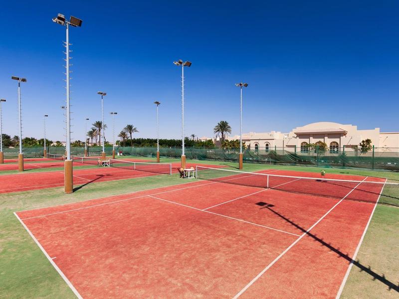 Red tennis courts with floodlights and blue sky on a hotel property.