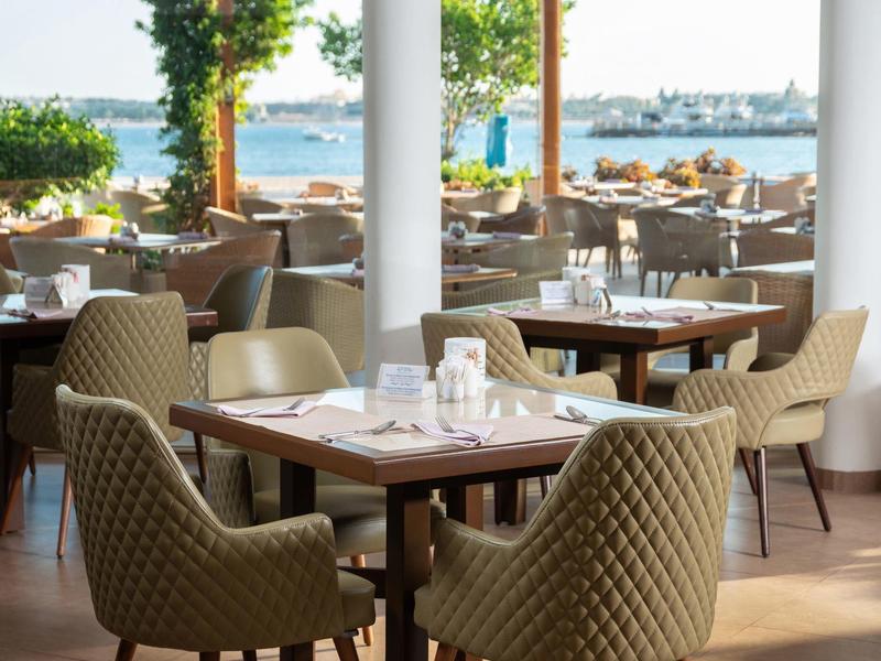 Outdoor restaurant seating with cushioned chairs overlooking the sea and distant ships.