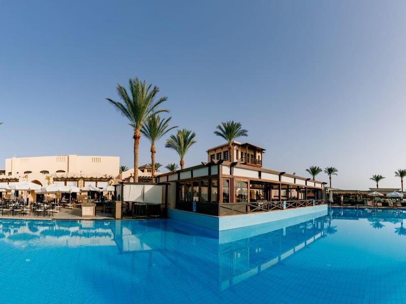 Piscine extérieure bleue claire avec palmiers et structure de détente sous un ciel dégagé.