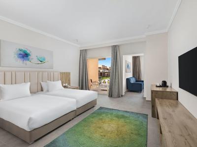 Helles Schlafzimmer mit großem Bett, grünem Teppich, TV an der Wand und Balkon mit Meerblick.