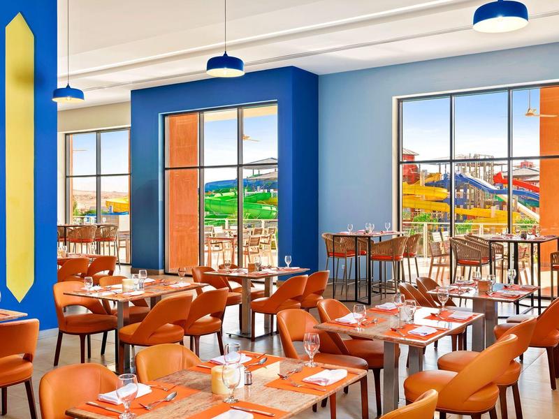 Helles Restaurant mit blauen Wänden, großen Fenstern und orangen Stühlen an Holztischen.