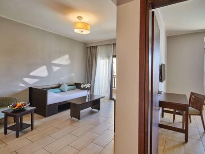 Jasny pokój hotelowy z częścią wypoczynkową, tapczanem, stołem do jadalni i dużymi oknami z zasłonami.