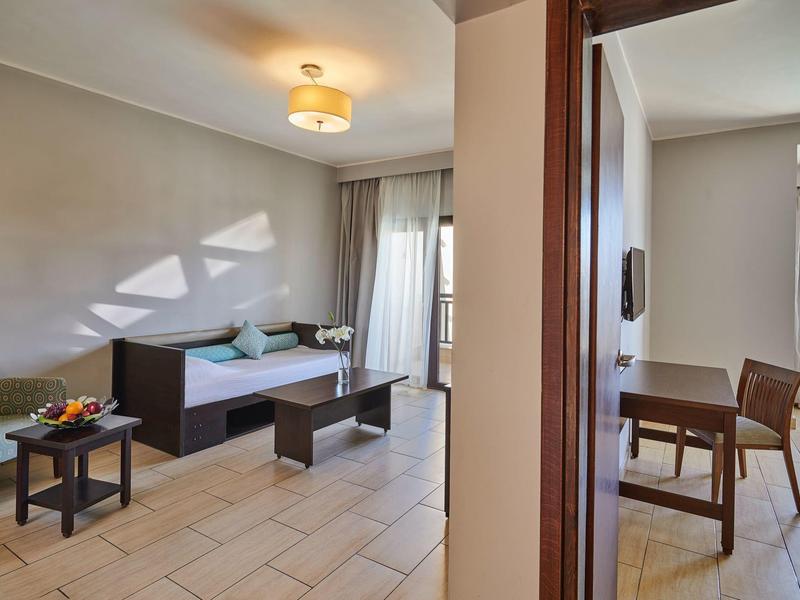 Jasny pokój hotelowy z częścią wypoczynkową, tapczanem, stołem do jadalni i dużymi oknami z zasłonami.