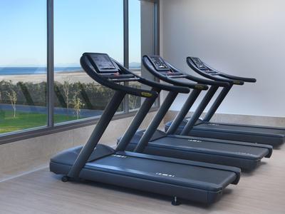 Vier Laufbänder in einem hellen Fitnessraum mit großen Fenstern und Meerblick.