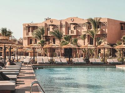 Hotel mit Pool, Sonnenschirmen und Liegestühlen vor einem mehrstöckigen Gebäude in sonniger Umgebung.