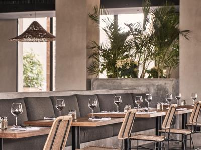 Bar d'hotel moderno con tavoli in legno, sedute imbottite grigie e piante vicino a grandi finestre.