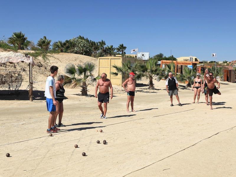 Persone che giocano a pétanque su un campo sabbioso sotto un cielo azzurro limpido con alberi e edifici sullo sfondo