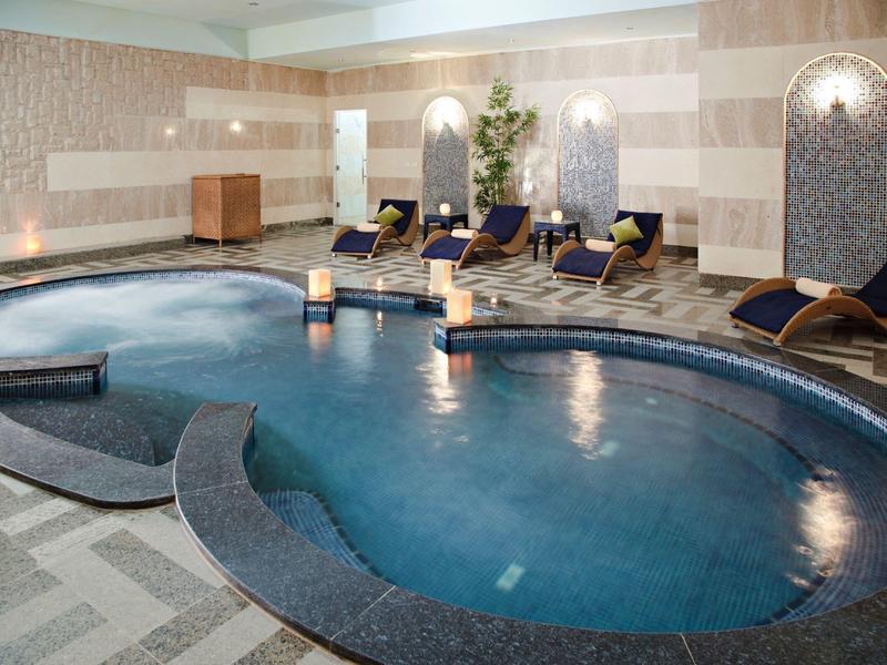 Piscina interna con acqua blu curva, lettini e luci calde alle pareti.