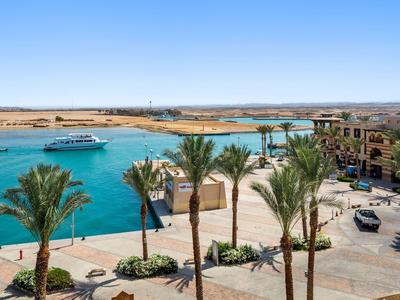 Paseo costero con palmeras, bahía azul, barco y edificios en un resort bajo cielo despejado.