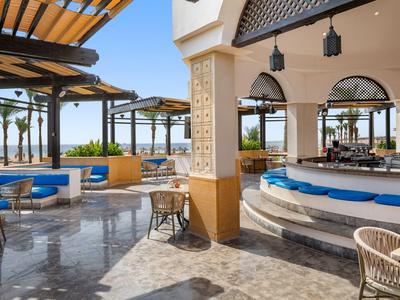 Zona lounge abierta con barra, asientos azules y vista al mar con clima soleado.